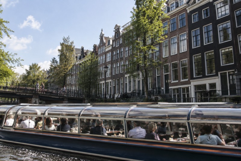 A low glass-roofed canal boat with passengers seated at small tables along the windows, drinks and snacks in front of them, gliding past tall narrow brick canal houses in Amsterdam