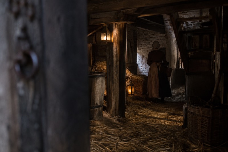 A dim theatrical cellar set with heavy timber beams, straw across the floor and lantern light, a costumed figure half lost in shadow, Amsterdam, Netherlands