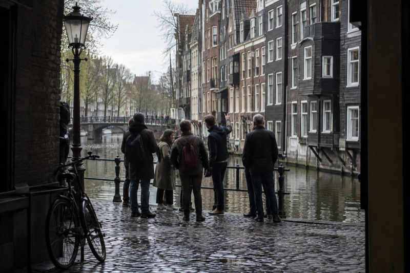 A small group standing with a guide beside the water in the oldest part of the city, timber-fronted houses leaning slightly over the canal behind them, Amsterdam, Netherlands