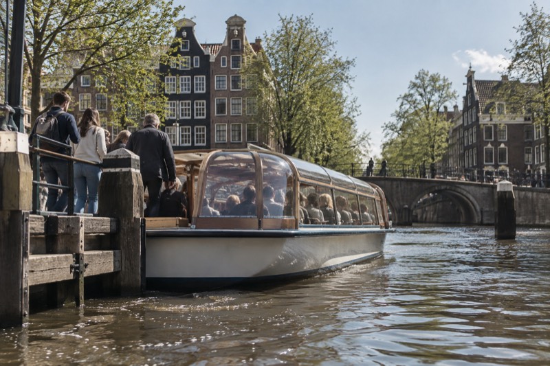 A glass-topped canal boat pulling away from a wooden mooring stage on a broad canal, passengers still stepping aboard from the quay, brick gables behind, Amsterdam, Netherlands