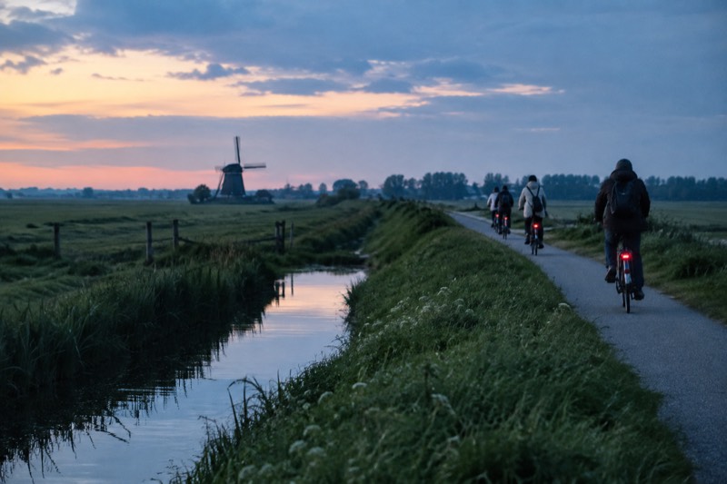 Cyclists strung out along a narrow raised path through flat green polder fields with a drainage ditch beside them and a windmill far off, countryside north of Amsterdam, Netherlands