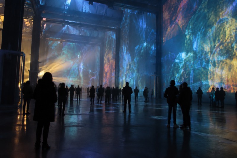 A darkened former industrial hall flooded with projected abstract colour and moving light, visitors standing as silhouettes on the open floor, Amsterdam, Netherlands