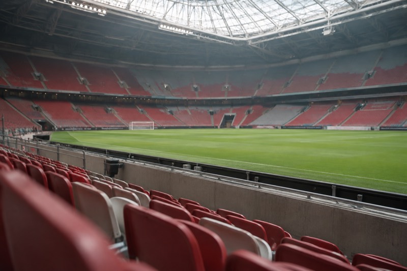 An empty modern football stadium seen from the lower tier, banks of red and grey seats rising around a bright green pitch under a closed roof, Amsterdam, Netherlands