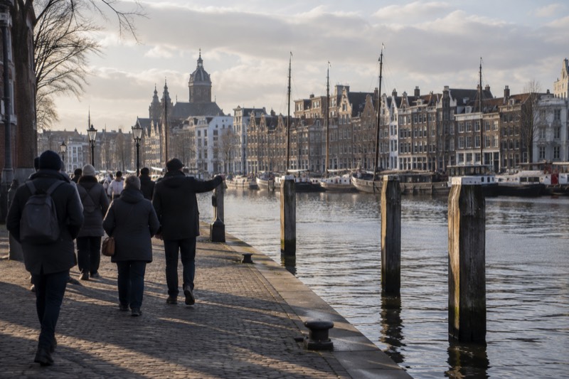 A guide and a handful of visitors on a broad harbour front with old timber mooring posts standing out of the water and a low brick quay, Amsterdam, Netherlands