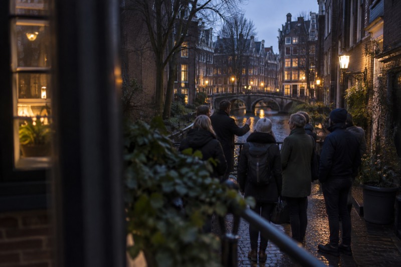 A guide addressing a small evening group on a narrow canal-side lane in the medieval quarter, warm light spilling from windows onto wet cobbles, Amsterdam, Netherlands