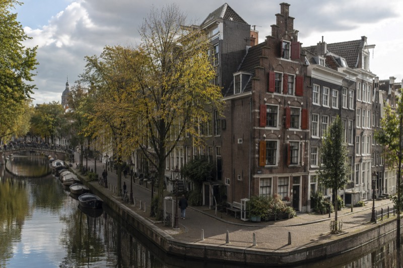 A tall seventeenth-century brick canal house with red-painted shutters and a stepped gable, narrow and pressed between its neighbours, seen from the street corner, Amsterdam, Netherlands