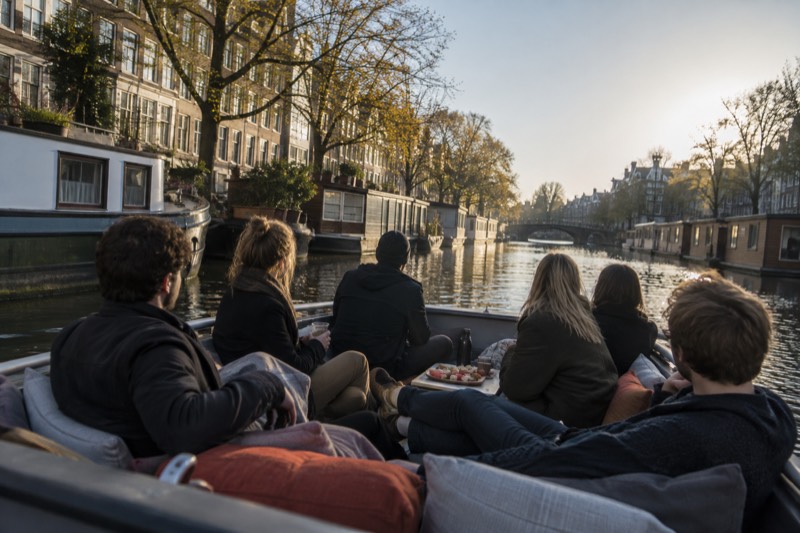 Six passengers on an open canal boat with cushions and a plate of snacks between them, passing a row of moored houseboats on a narrow canal, Amsterdam, Netherlands
