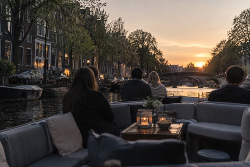 A softly lit lounge boat with deep cushioned benches and low tables on a canal at dusk, a few passengers seated with their backs to the camera, Amsterdam, Netherlands