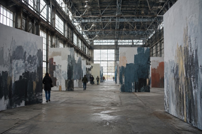 The interior of an enormous former shipbuilding warehouse with tall steel roof trusses and daylight from high windows, large painted panels standing in rows, visitors small in the space