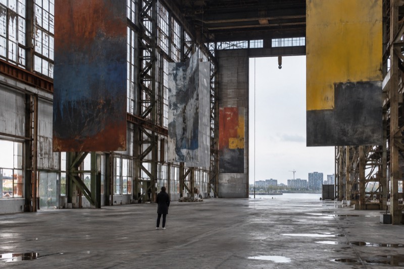 A towering steel-framed industrial hall on a former shipyard, abstract colour on hanging metal panels and a raw concrete floor, a single visitor looking up, Amsterdam Noord, Netherlands