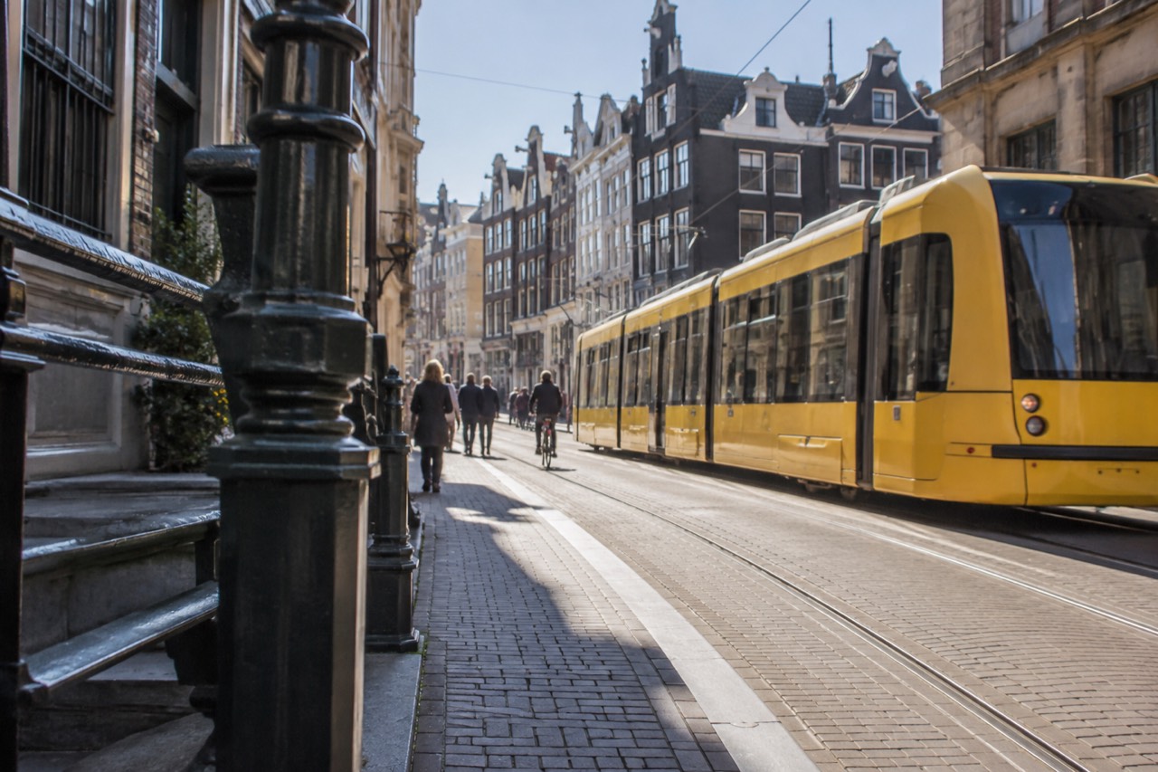 A yellow Amsterdam tram passing gabled canal houses on a narrow street