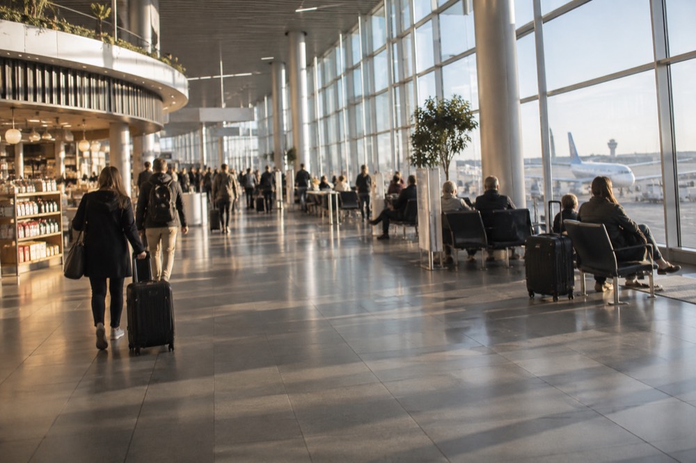 A bright airport terminal concourse with shops and seated travellers beside tall windows