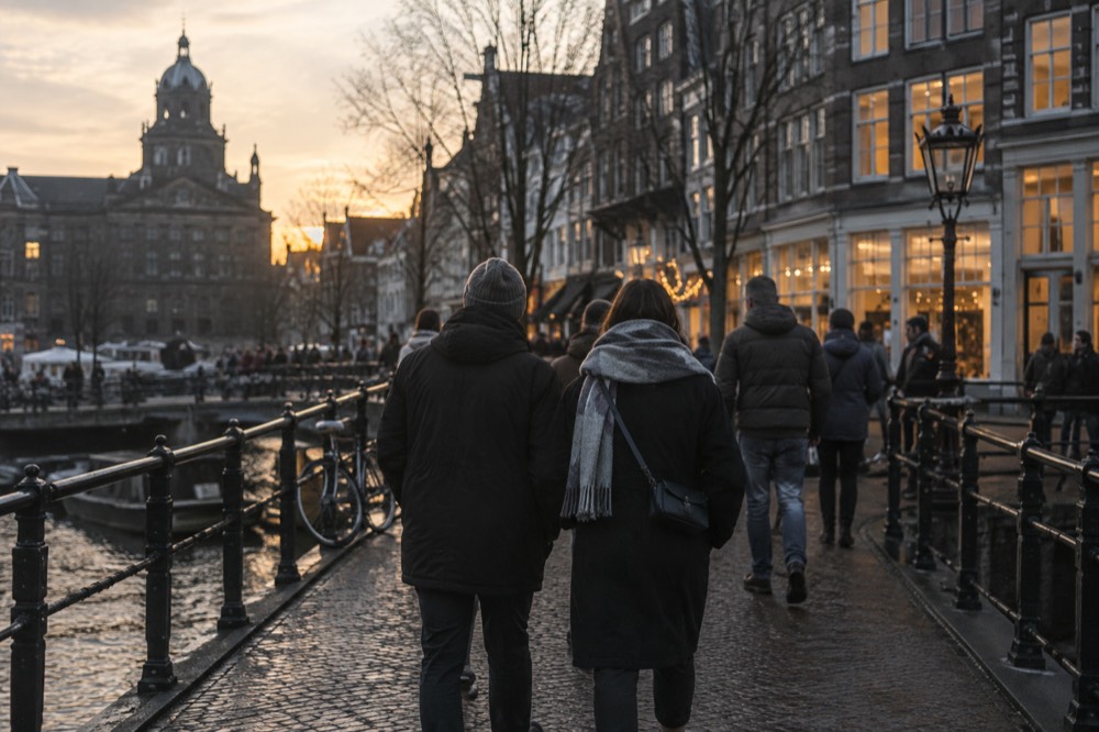 Winter visitors walking across a small Amsterdam bridge towards lit shopfronts in the early evening