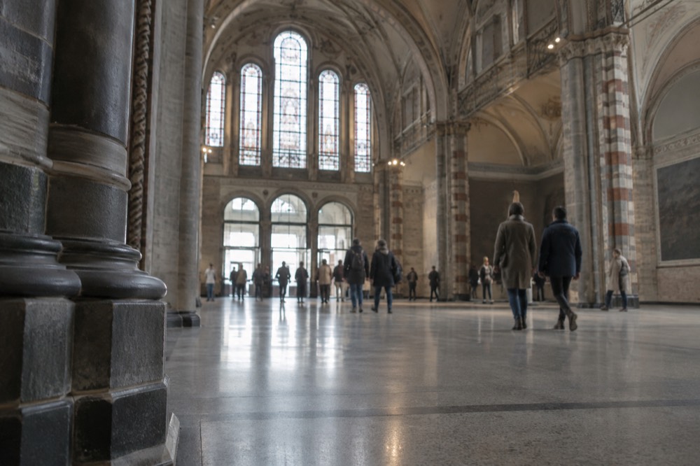 The grand arched entrance hall of the Rijksmuseum with stained glass windows and visitors walking across the polished stone floor