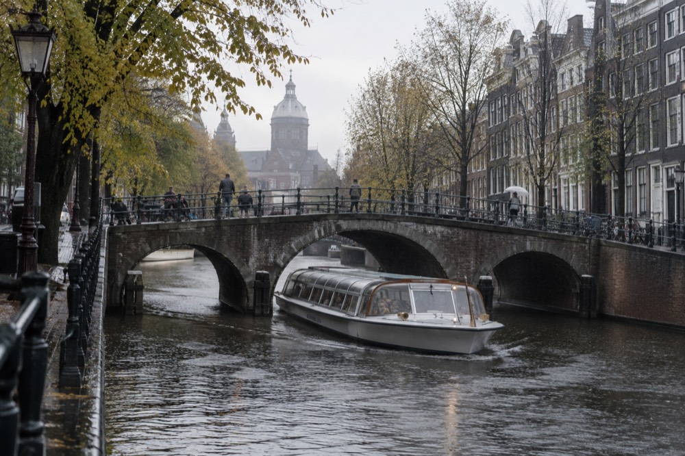 A covered canal cruise boat passing under a stone bridge in Amsterdam in soft rain