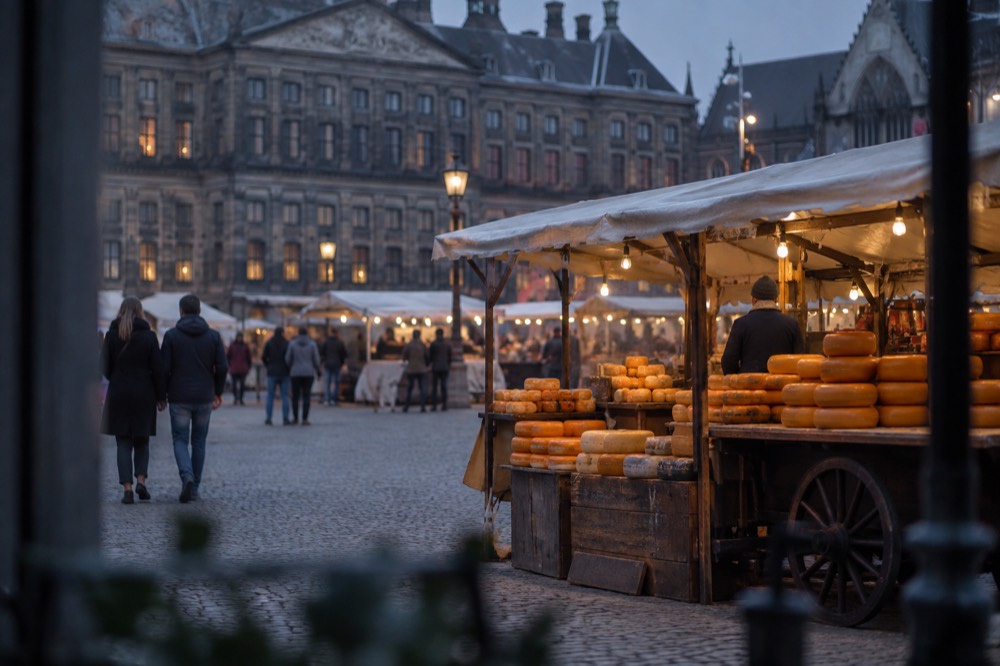 A market stall with wheels of Dutch cheese stacked under a canvas awning