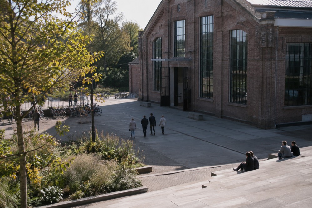 A tall brick industrial hall with large windows and a paved courtyard in front
