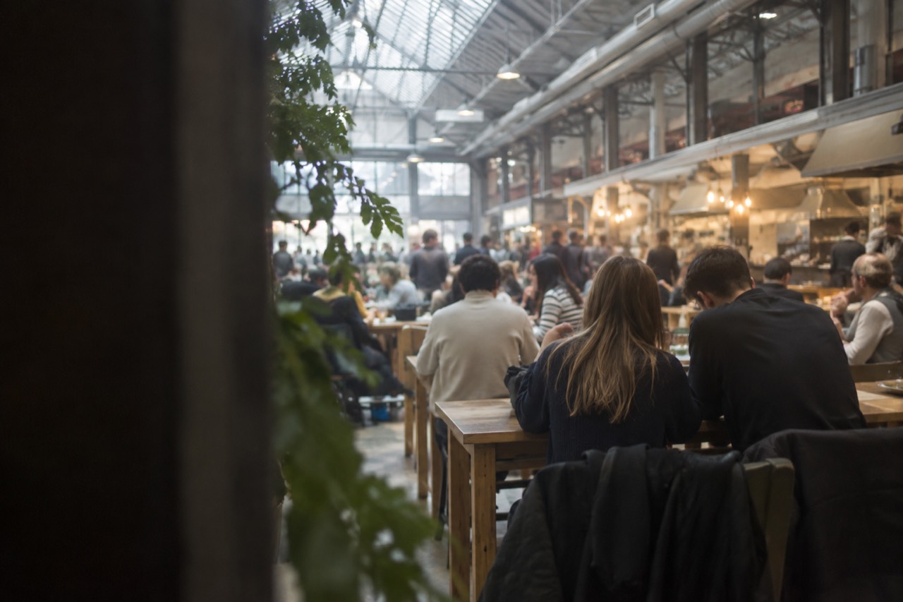 Interior of a busy food hall with food stalls and shared wooden tables under a glass roof