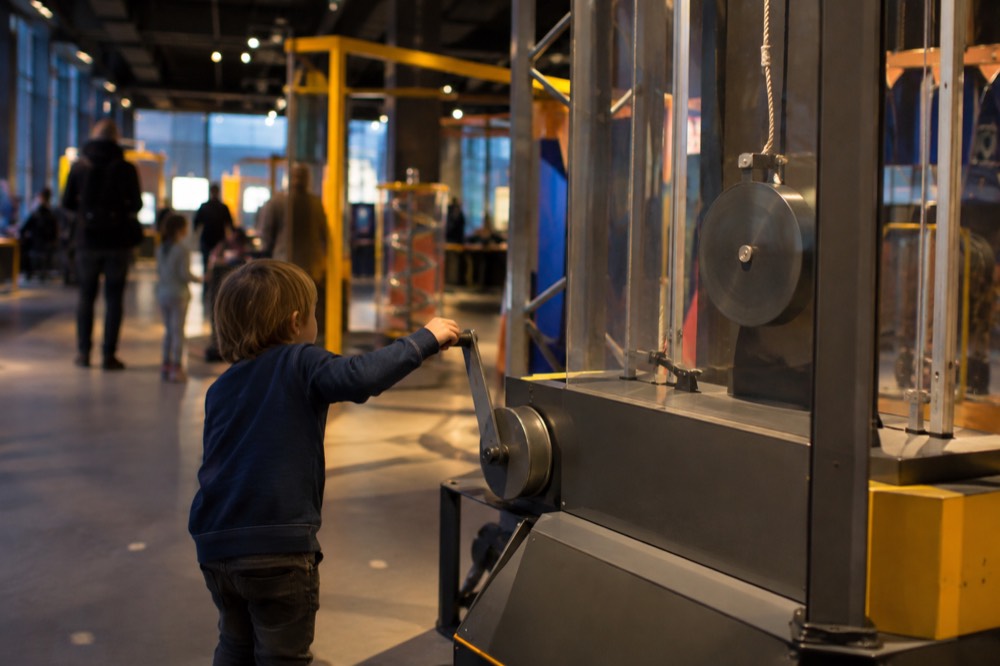 Inside the NEMO Science Museum, a child pulls a lever on a large interactive physics exhibit while coloured lights flash