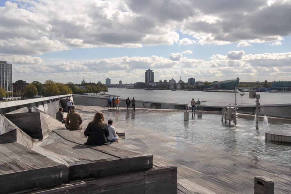 The rooftop terrace of NEMO Science Museum with large outdoor water exhibits and visitors looking toward Amsterdam's city centre under a blue sky