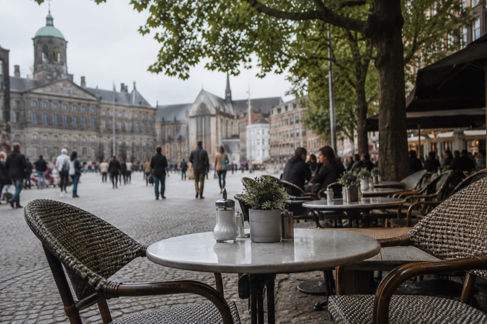 Cafe terraces with chairs and small tables under trees along the edge of a city square in late afternoon light