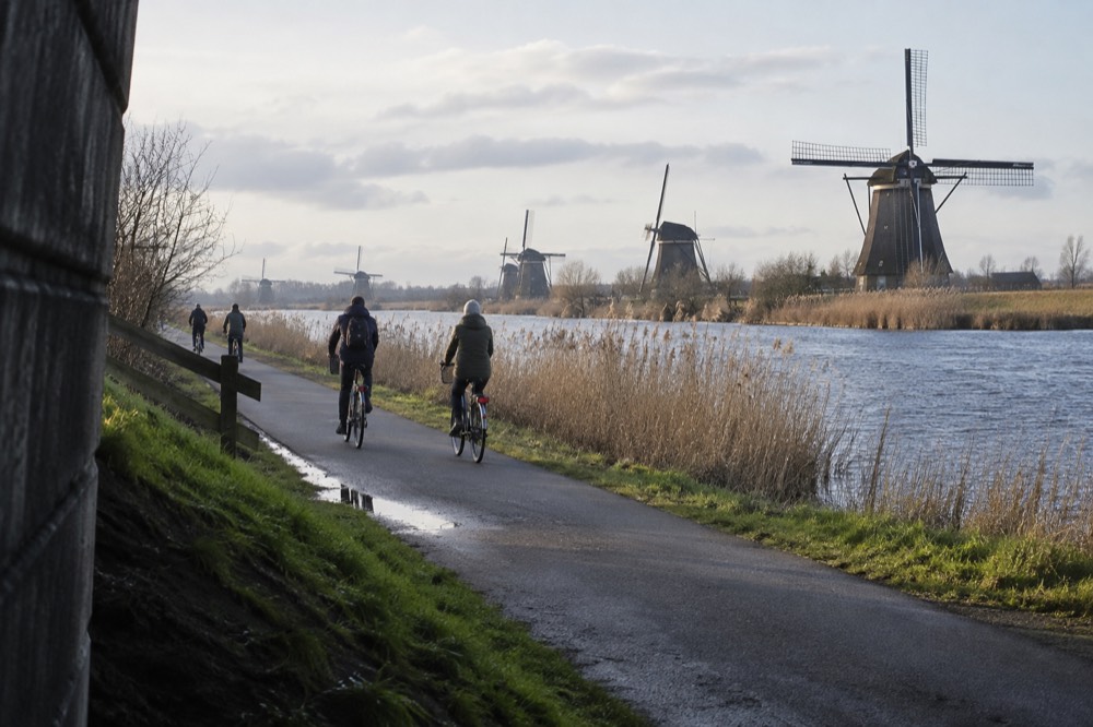 Cyclists on a path beside a canal with windmills in the background