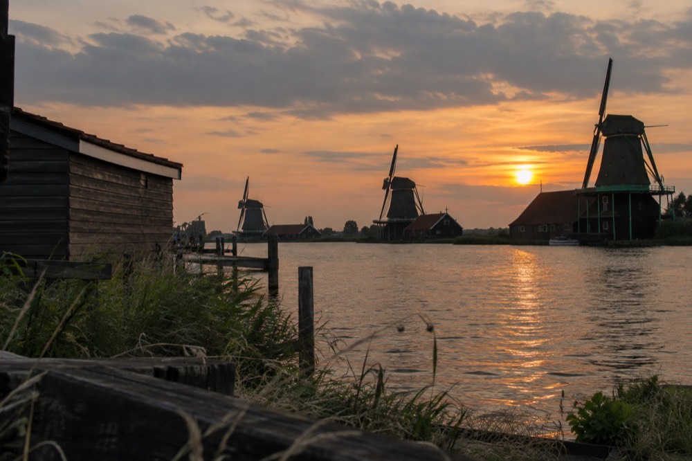 Wide view of windmills along a river at dusk with dark wooden silhouettes against an orange sky