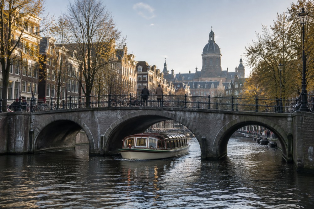 A covered saloon boat gliding under a stone canal bridge with autumn trees along the bank