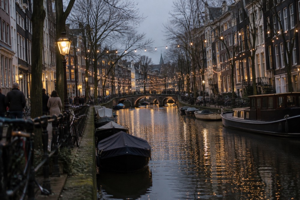 Warm white fairy lights strung across a narrow Amsterdam canal at dusk with boats moored below