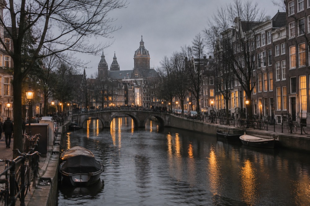 A quiet Amsterdam canal at dusk with warm light reflecting on the water and bare trees along the quay