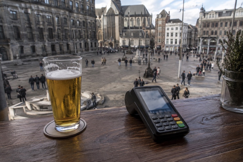 A contactless payment terminal sitting on a wooden bar counter next to a glass of Dutch beer