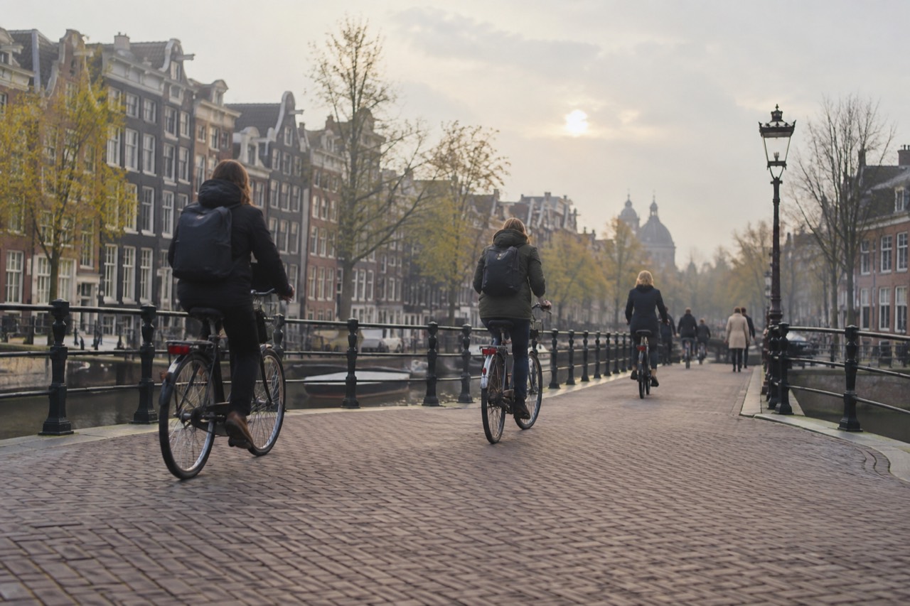 Cyclists in jackets crossing a canal bridge lined with narrow brick houses