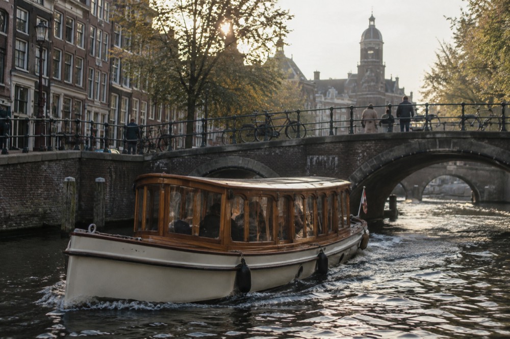 A wooden saloon boat passing under a bridge on an Amsterdam canal in late afternoon light