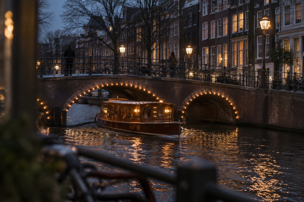 A classic wooden saloon boat passing under a lit bridge on an Amsterdam canal at night